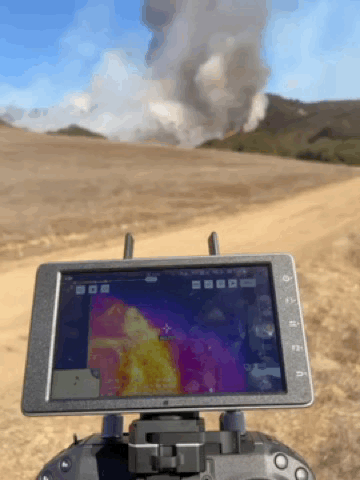 Animation: a drone controller screen showing a live thermal view of the fire, with the real smoke plume rising from the hillside behind it